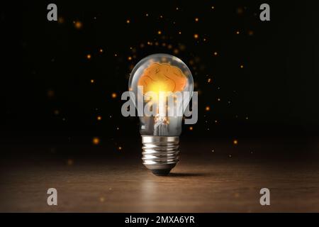 Glühlampe mit glänzendem Gehirn auf einem Holztisch vor schwarzem Hintergrund. Ideengenerierung Stockfoto