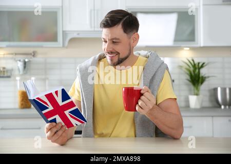 Ein hübscher Mann mit einer Tasse Kaffee, der ein Buch am Tisch in der Küche liest. Englisch Lernen Stockfoto