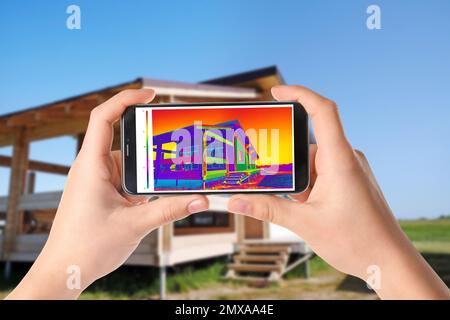 Frau erkennt Wärmeverlust im Haus mit Thermokino auf Smartphone und im Freien. Energieeffizienz Stockfoto