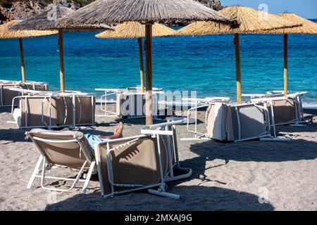 Umgedrehte Sonnenschirme und Sonnenliegen am Strand, Kreta, Griechenland Stockfoto