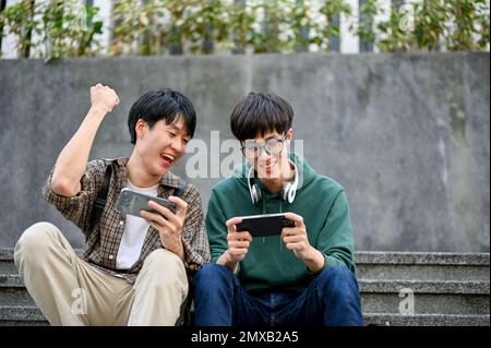 Fröhlich und überglücklich junger asiatischer männlicher Student, der mit seinem Freund ein mobiles Spiel spielt, seinen Freund anfeuert und Spaß zusammen hat. Stockfoto