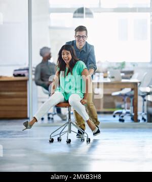 Eins, zwei, drei. LOS. Ein männlicher Mitarbeiter, der seine weibliche Mitarbeiterin in einem Stuhl durch ein Büro schubst. Stockfoto