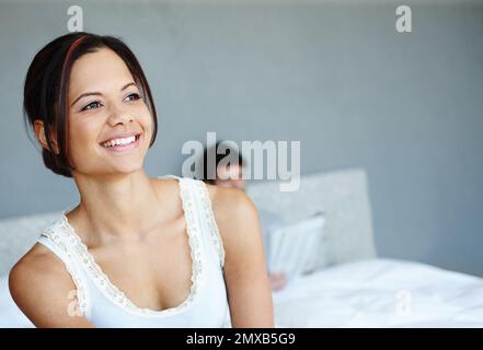 Entspannt und erfrischt. Eine lächelnde Frau auf der Seite ihres Bettes mit ihrem Freund im Hintergrund. Stockfoto
