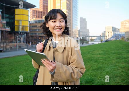 Glückliches asiatisches Mädchen steht auf der Straße, Universitätsstudentin läuft mit einem digitalen Tablet in den Händen und lächelt, steht im Stadtzentrum Stockfoto