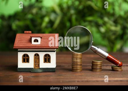 Hausmodell, Münzen und Lupe auf Holztisch vor verschwommenem Hintergrund. Suchkonzept Stockfoto
