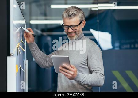 Ein erwachsener Mann im Büro neben dem Whiteboard mit farbigen Notizen, die Strategie und Geschäftsplan aufschreiben, ein grauhaariger Geschäftsmann mit Tablet-Gedanken bei der Arbeit im Büro, der mit dem Ergebnis zufrieden lächelt. Stockfoto