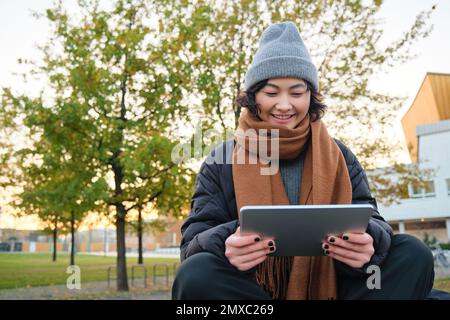 Porträt eines asiatischen Mädchens in warmer Kleidung, sitzt auf einer Bank mit digitalem Tablet und Stift, lächelt fröhlich und zieht bei kaltem Wetter ins Freie Stockfoto