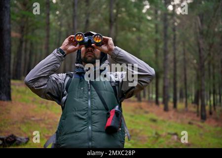 Vogelbeobachter auf der Jagd. Ein gutaussehender Mann, der sein Fernglas benutzt, um den umliegenden Wald nach Vögeln zu scannen. Stockfoto