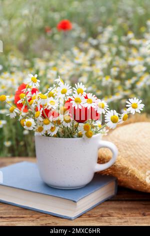 Komposition mit Kamillen, Mohn, Strohhut und Buch auf Holztisch im Freien Stockfoto