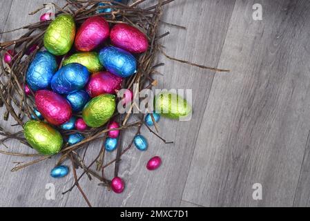 In Folie gewickelte mehrfarbige ostereier in rosa, grün, blau und gelb in einem natürlichen Nest aus Stäben und Zweigen, gegen einen grau weißen Holzhinterholz Stockfoto