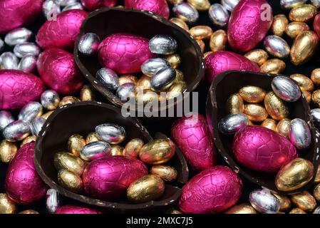 Ein Stapel bunter und in verschiedenen Größen mit bunten Folien verpackte Schokoladeneier in Rosa, Rot, Silber und Gold mit zwei Hälften eines großen Eies. Stockfoto