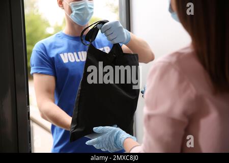 Männlicher Freiwilliger gibt der Frau schwarze Tasche durch die Tür, schließen. Hilfe während der Coronavirus-Quarantäne Stockfoto