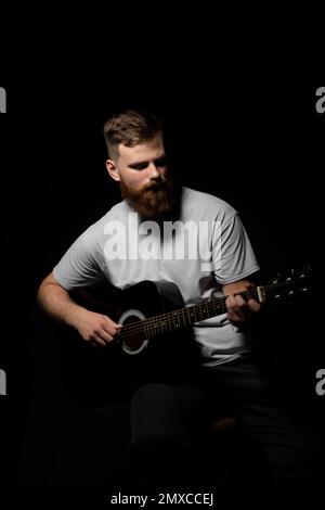 Porträt eines gutaussehenden jungen bärtigen Musikers, Gitarrenspieler spielt ein Lied mit einer Akustikgitarre in einem dunklen Raum. Stockfoto