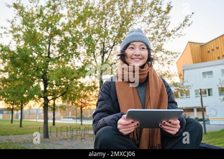 Porträt eines asiatischen Mädchens in warmer Kleidung, sitzt auf einer Bank mit digitalem Tablet und Stift, lächelt fröhlich und zieht bei kaltem Wetter ins Freie Stockfoto