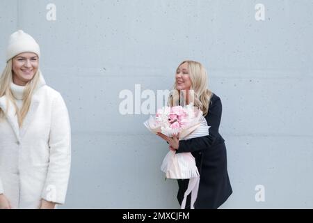 Eine überfreuliche Frau im Herbstmantel mit einem wunderschönen Blumenstrauß an der grauen Wand. Nette Kurierfrau liefert den Auftrag. Stockfoto