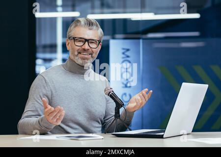 Erfolgreicher, erfahrener Finanzberater, der im Büro mit Laptop und professionellem Mikrofon arbeitet, Senioren, die Podcasts aufzeichnen und Hörbuchmentoren, die in die Kamera schauen und lächeln. Stockfoto
