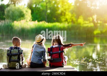 Schulferien. Gruppe von Kindern, die auf einem hölzernen Pier in der Nähe des Flusses sitzen Stockfoto