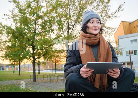 Porträt eines asiatischen Mädchens in warmer Kleidung, sitzt auf einer Bank mit digitalem Tablet und Stift, lächelt fröhlich und zieht bei kaltem Wetter ins Freie Stockfoto