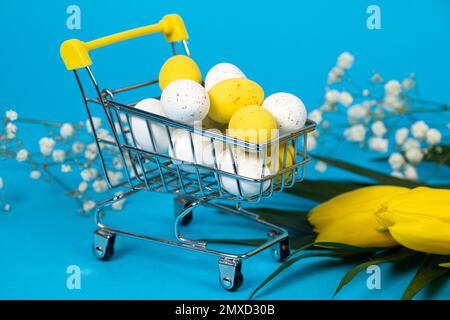 Osterweißes und gelbes Wachteln bemalte Eier in einem Einkaufswagen auf blauem Hintergrund. Osterangebote und -Shopping Stockfoto