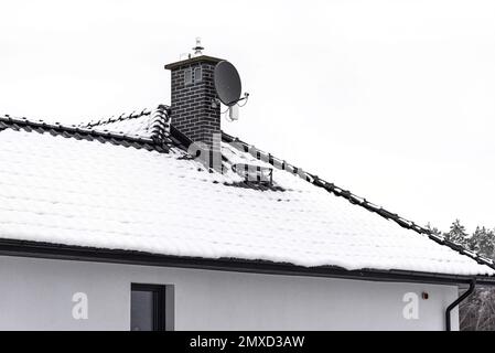 Das Dach eines Einfamilienhauses ist mit Schnee bedeckt vor einem bewölkten Himmel, einem sichtbaren Keramikkamin und einer Satellitenschüssel. Stockfoto