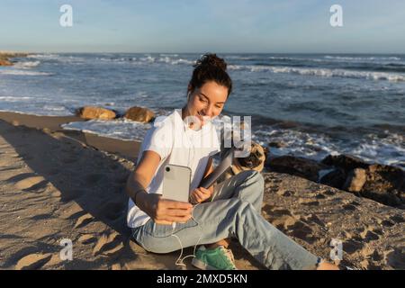 Fröhliche Frau in kabelgebundenen Kopfhörern, die einen Laptop in der Hand hält und in Spanien ein Selfie mit einem Hund am Meer macht Stockfoto