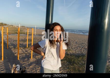 Fröhliche junge Frau, die mit dem Smartphone spricht, während sie am Strand von Barcelona mit der Hand auf der Hüfte steht Stockfoto
