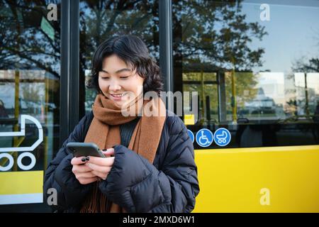 Junge, schöne Frau, die an der Bushaltestelle steht, SMS auf dem Smartphone schreibt, ihr Handy hält, ihren Terminplan überprüft, Fahrkarte online kauft, trägt Stockfoto