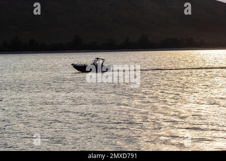 Ein Motorboot fährt mit hoher Geschwindigkeit auf dem Wasser. See mit felsigen Ufern, abendliche Landschaft. Stockfoto