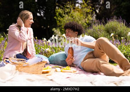 Glückliches, vielfältiges Paar, das sich auf einer Decke entspannt und ein Picknick im sonnigen Garten macht Stockfoto