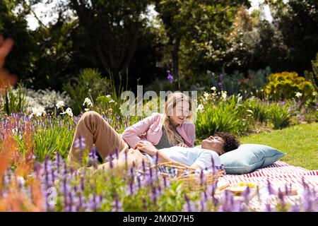 Glückliches, vielseitiges Paar, das auf einer Decke lag, ein Picknick machte, sich im sonnigen Garten unterhielt Stockfoto