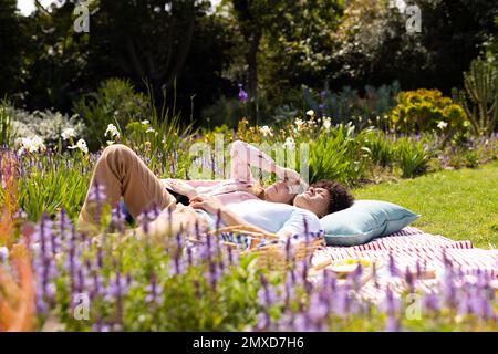 Glückliches, vielseitiges Paar, das auf einer Decke lag und sich im sonnigen Garten unterhielt Stockfoto