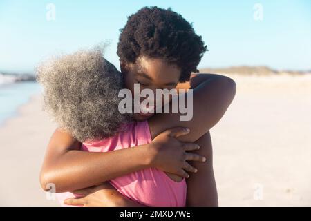 Rückansicht einer afroamerikanischen Seniorin, die eine glückliche junge Tochter am Strand unter blauem Himmel umarmt Stockfoto