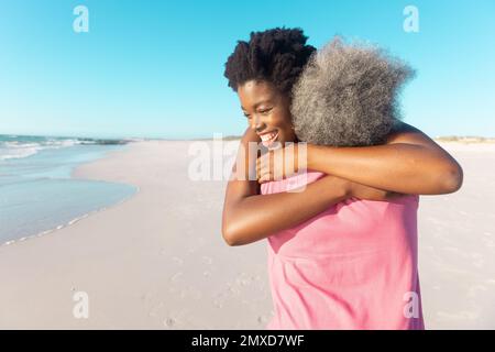 Rückblick auf die afroamerikanische Seniormutter, die eine glückliche junge Tochter am Strand unter blauem Himmel umarmt Stockfoto