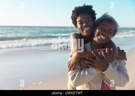 Fröhliche junge afroamerikanische Tochter, die ältere Mutter umarmt, während sie sich gegen Meereslandschaft stellt Stockfoto