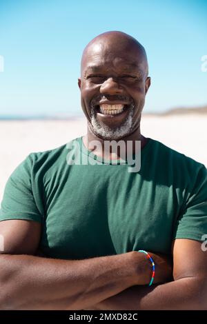 Sorgloser, kahlköpfiger, afroamerikanischer Seniorist mit gekreuzten Armen, der am Strand vor klarem Himmel lächelt Stockfoto