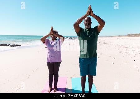 afroamerikanisches Seniorenpaar, das in den Bergen meditiert, posiert am Strand vor klarem Himmel Stockfoto