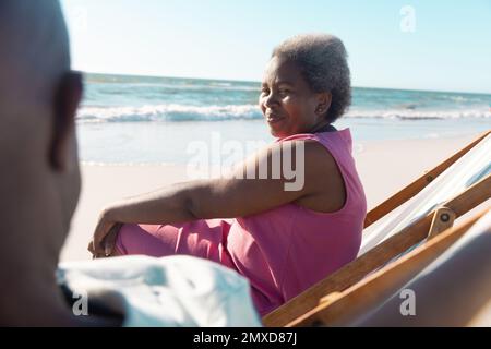 Lächelnde afroamerikanische Seniorin, die sich mit einem Mann unterhielt, während sie sich am Strand auf Liegestühlen entspannte Stockfoto