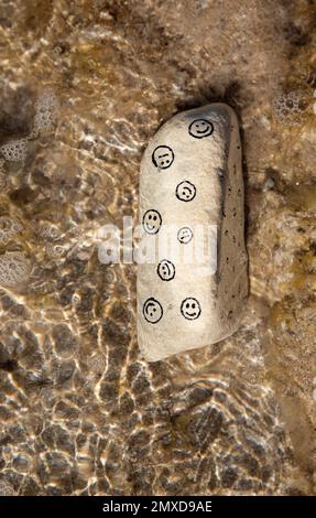 Weißer Stein mit lustigen und traurigen Maulkörben auf einem Sandstrand. Sonnenlicht im Meerwasser. Hallo Sommer, Sonne, Urlaub, Reisen. Stimmungsauswahl, BE Stockfoto