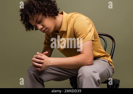 Junger afroamerikanischer Mann mit lockigem Haar, trägt ein gelbes Poloshirt und sitzt auf einem Stuhl mit geballten Händen isoliert auf grauem Stockbild Stockfoto