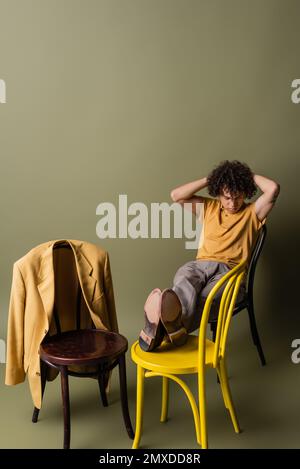 Ein eleganter afroamerikanischer Typ in voller Länge, mit Händen hinter dem Kopf, auf schwarzen und gelben Stühlen auf graugrünem Hintergrund, Bild von der Börse Stockfoto