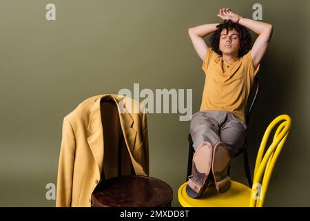 Junger afroamerikanischer Mann mit geschlossenen Augen und Händen hinter dem Kopf, sitzt auf Stühlen neben einem trendigen gelben Blazer auf graugrünem Hintergrund, Stockbild Stockfoto