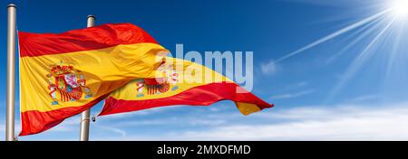 Nahaufnahme von zwei spanischen Nationalflaggen (La Rojigualda) mit Fahnenstange, die im Wind an einem blauen Himmel mit Wolken, Sonnenstrahlen und Kopierraum weht. Stockfoto