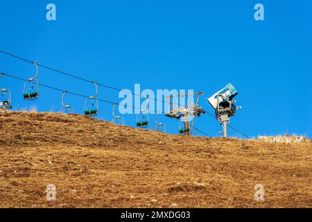 Schneekanone oder Schneemaschine und ein leerer Sessellift im Winter auf einer braunen Wiese, Skipiste, ohne Schnee wegen der zu Hitze. Veneto, Italien. Stockfoto