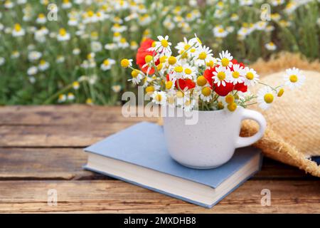 Komposition mit Kamillen, Mohn, Strohhut und Buch auf Holztisch im Freien Stockfoto