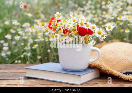 Komposition mit Kamillen, Mohn, Strohhut und Buch auf Holztisch im Freien Stockfoto