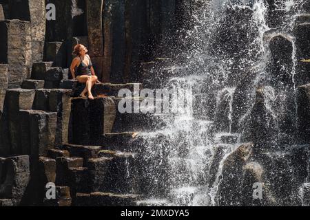 Eine weiße Frau in schwarzem Badeanzug, die die Wasserläufe in schwarzen Vulkansteinkaskaden genießt. Rochester Falls Wasserfall - beliebter tou Stockfoto