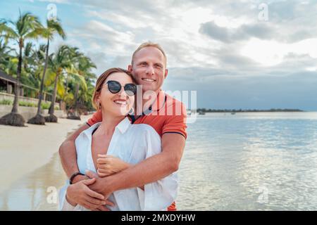 Lächelndes Paar, das sich am exotischen Sandstrand umarmt, während Sie einen Abendspaziergang an der Küste von Trou-aux-Biches auf Mauritius machen. Beziehungen zu Menschen Stockfoto