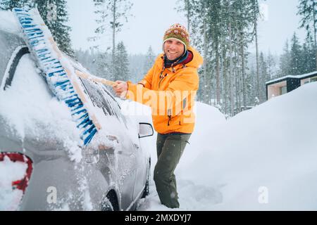 Ein lächelnder Mann in einem lustigen nepalesischen Hut, der Schnee mit einer großen Schneebürste aus dem Auto entfernt. Das Auto friert auf der Landstraße des Waldhauses. Gewinnen Stockfoto