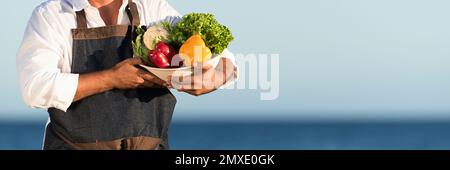 Der Mann hält eine Schüssel mit frischem Gemüse, blauer Himmel im Hintergrund Stockfoto