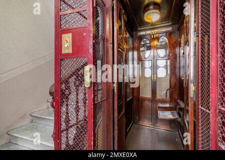 Leerer Aufzug im Retro-Stil mit roten Metallstangen, Türen auf beiden Seiten, innen mit dunkel polierten Holzwänden. Der Fahrstuhl wird von einer Lampe mit einem beleuchtet Stockfoto
